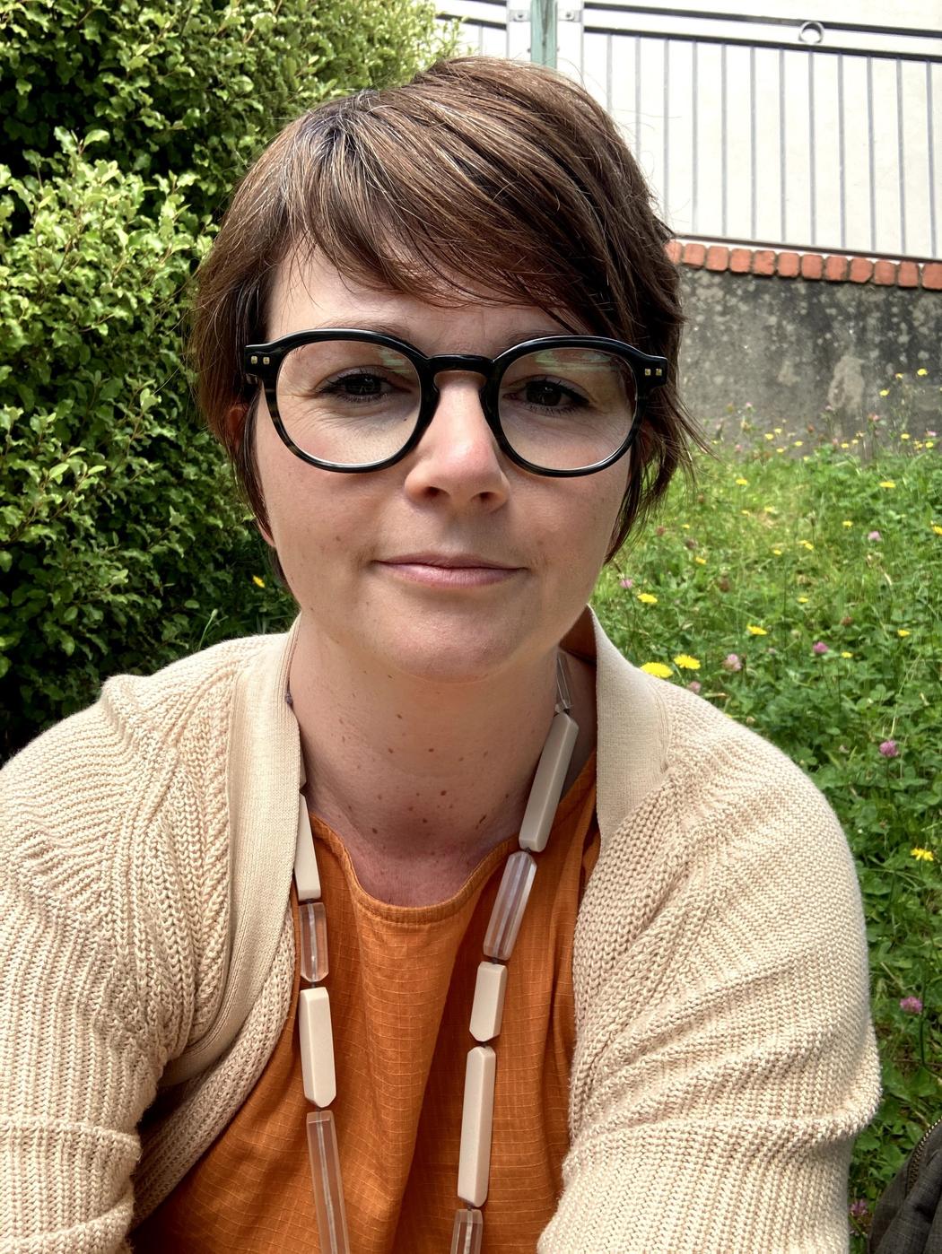 A person looking at the camera wearing glasses and an orange top